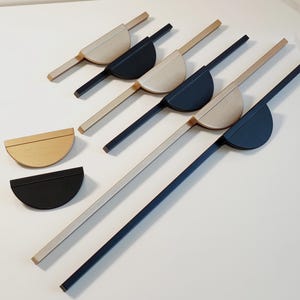 May include: A collection of modern cabinet pulls in various colors. The pulls feature a half-circle design with a long, rectangular handle. Colors include natural wood, black, and off-white. The hardware is arranged on a white surface.
