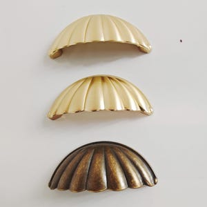 May include: Three shell-shaped cabinet pulls in gold and bronze. The top two are bright gold, the bottom one is antique bronze. Each pull has a ribbed, fan-like design. Suitable for drawers or cupboards. Measurements are in centimetres.