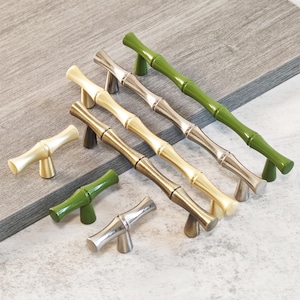 Puede incluir: Un conjunto de tiradores de armario con diseño de bambú en varios colores, incluyendo verde, plateado, dorado y bronce. Los tiradores están dispuestos sobre una superficie gris claro, mostrando su diseño decorativo y su potencial para proyectos de decoración del hogar.
