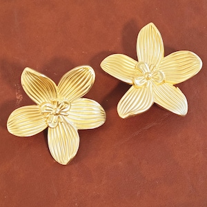 Op de afbeelding: Twee goudkleurige bloemvormige decoratieve objecten. Elke bloem heeft vijf bloemblaadjes met gedetailleerde nerven en een centrale, getextureerde kern. De objecten zijn waarschijnlijk van metaal en staan tegen een bruine achtergrond.