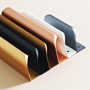 May include: A row of metal shelf brackets in various colors: gold, black, copper, white, and gray. Each bracket has a right-angle design with a flat base and a curved top, suitable for supporting shelves or other items.