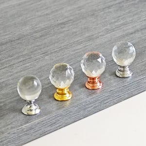 Könnte beinhalten: Vier dekorative Kristallkugel-Schrankknöpfe mit facettierten Oberflächen. Die Knöpfe haben verschiedene Grundfarben: Silber, Gold, Kupfer und Silber. Die Knöpfe werden auf einer grauen Holzoberfläche präsentiert.