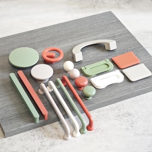 Puede incluir: Una colección de herrajes para armarios en varias formas y colores, incluyendo pomos, tiradores y manijas. Los herrajes son en tonos de verde, rojo, blanco y gris. Los artículos están dispuestos sobre una superficie de madera gris.