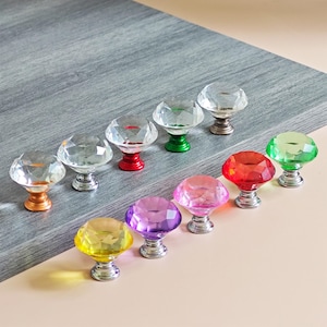 Könnte beinhalten: Eine Sammlung von zehn facettierten Kristall-Schrankknöpfen in verschiedenen Farben, darunter klar, gold, rot, grün, lila und gelb. Jeder Knopf hat einen silbernen Sockel und ist diagonal angeordnet.