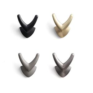 May include: Four V-shaped hooks in different finishes: black, gold, nickel, and brushed nickel. The hooks are designed for hanging items on a wall.