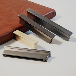 May include: A selection of cabinet handles in various colours. There are three dark grey metal handles and one cream-coloured handle. The handles are rectangular with a beveled design. A brown leather-bound notebook is in the background.