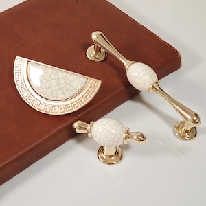 Może przedstawiać: Zestaw złotych okuć do szafek, w tym uchwyt w kształcie półksiężyca z pękniętym białym środkiem i obramowaniem w grecki wzór, a także dwa gałki i uchwyt z białymi ceramicznymi akcentami z efektem spękania. Na brązowej powierzchni.