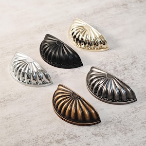 May include: Five decorative half-moon cabinet pulls in various metallic finishes: silver, black, gold, bronze, and pewter. Each pull features a ribbed, fan-like design. These hardware pieces are ideal for updating kitchen or bathroom cabinets.