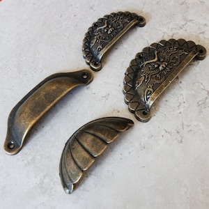 May include: Four antique-style cabinet pulls in various designs. Two are half-circle shaped with ornate details, one is a simple half-circle, and one is a scallop-shaped pull.
