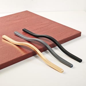 May include: Four curved cabinet handles in gold, silver, gray, and black. The handles are placed on a reddish-brown wooden surface. The handles have a modern, minimalist design.