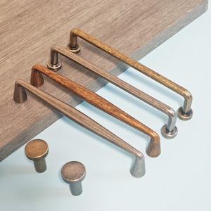 Op de afbeelding: Een assortiment kastbeslag, waaronder vier lange, rechthoekige handgrepen en twee ronde knoppen. De handgrepen variëren in kleur, van brons tot zilver, en zijn gerangschikt op een lichtblauwe ondergrond. De knoppen zijn bronskleurig.