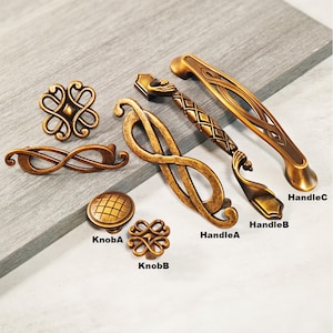 Może przedstawiać: Kolekcja antycznych mosiężnych okuć do szafek, w tym gałek i uchwytów. Okucia charakteryzują się skomplikowanymi wzorami, z etykietami identyfikującymi je jako KnobA, KnobB, HandleA, HandleB i HandleC. Okucia są prezentowane na szarej powierzchni.