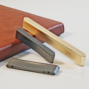 Peut inclure: Trois poignées de meubles modernes en or, gris foncé et gris clair. La poignée dorée est inclinée, tandis que les deux autres sont droites. Un article en cuir marron est en arrière-plan. Les poignées sont rectangulaires avec des bords biseautés.
