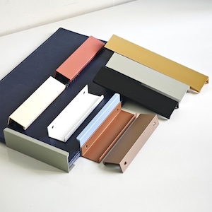 Könnte beinhalten: Eine Sammlung farbenfroher, moderner Schrankgriffe in verschiedenen Farbtönen, darunter Weiß, Schwarz, Silber, Gold und Blau. Die Griffe sind rechteckig mit einer gebogenen Lippe und auf einer dunkelblauen Oberfläche angeordnet.