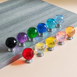 Op de afbeelding: Een verzameling kleurrijke kristallen kastknoppen. De knoppen zijn facetgeslepen en bolvormig, met een verscheidenheid aan kleuren, waaronder zwart, paars, groen, blauw, turquoise, geel, roze en oranje. Elke knop is gemonteerd op een zilverkleurige basis.