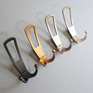 Könnte beinhalten: Vier dekorative Metall-Wandhaken in verschiedenen Metallic-Ausführungen: Schwarz, Bronze, Silber und Gold. Jeder Haken hat einen rechteckigen oberen Teil mit einem Ausschnitt und einen gebogenen Haken mit einem dekorativen Kugelende. Die Haken sind diagonal vor grauem Hintergrund angeordnet.