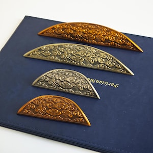 Könnte beinhalten: Vier dekorative Metall-Schubladengriffe in Bronze- und Silbertönen. Jeder Griff hat eine halbkreisförmige Form mit einem wolkenartigen Muster. Die Griffe sind auf einer dunkelblauen Oberfläche angeordnet.