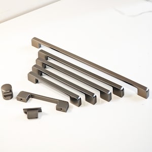 以下が含まれることがあります： ガンメタルカラーのキャビネットハードウェアのコレクション。さまざまなサイズのバープル、小さなノブ、長方形のプルが含まれています。 ハードウェアは白い表面に配置されており、さまざまなデザインとサイズが示されています。