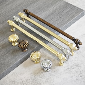 May include: A collection of cabinet hardware, including gold, silver, and bronze-toned pulls and knobs. The pulls are long and slender, while the knobs have a flower-like design. The hardware is arranged on a gray surface, showcasing various finishes and styles.