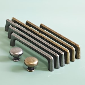 May include: A collection of cabinet hardware, including several metal bar pulls and two round knobs. The pulls are in various finishes, including silver, bronze, and antique brass. The knobs are also metallic, with a similar aged appearance.