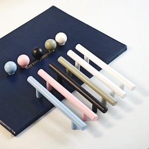 Könnte beinhalten: Eine Auswahl an Schrankgriffen und -knöpfen in verschiedenen Farben. Die Knöpfe sind hellblau, rosa, schwarz, olivgrün und weiß. Die Stangengriffe sind hellblau, rosa, schwarz, olivgrün und weiß. Auf einer dunkelblauen Oberfläche angeordnet.