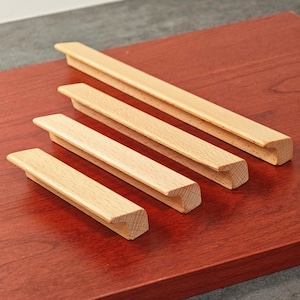 May include: Four wooden cabinet pulls of varying lengths, arranged on a reddish-brown surface. The pulls are rectangular with a recessed grip area and a slightly angled top edge. The wood has a light, natural finish.