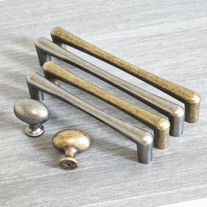 Op de afbeelding: Een verzameling kastbeslag, waaronder vier lange stangtrekkers en twee knoppen. De trekkers hebben een licht gebogen ontwerp en zijn afgewerkt in antiek messing en zilverkleurige tinten. De knoppen zijn ovaal van vorm en hebben een vergelijkbare afwerking.