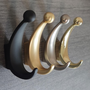 Może przedstawiać: Cztery dekoracyjne haczyki ścienne w rzędzie, każdy o zakrzywionym kształcie i kulką na górze. Haczyki są w kolorach matowej czerni, złota, srebra i błyszczącego złota. Zamontowane na drewnianej podstawie na szarej ścianie.