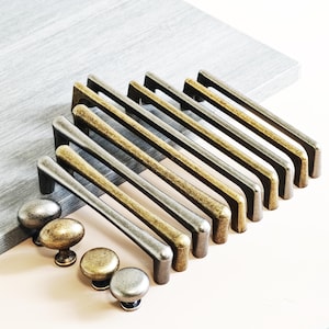 Peut inclure: Ensemble de poignées et boutons de meubles métalliques. L'image présente des tirettes rectangulaires argentées, dorées et bronze, ainsi que des boutons ronds aux finitions similaires. L'ensemble est disposé sur une surface claire, mettant en valeur les différents styles.