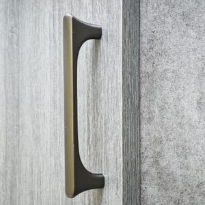 以下が含まれることがあります： 暗い茶色のアクセントが付いたブロンズ色のキャビネットプルハンドル。ハンドルは、明るい灰色の木製キャビネットドアに取り付けられています。ハンドルは、湾曲した端を持つ長方形の形状をしています。