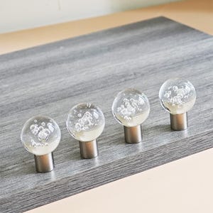 Könnte beinhalten: Vier transparente Glasschrankknöpfe mit einem Blasendesign und silberfarbenen Metallsockeln. Die Knöpfe sind in einer Reihe auf einer grauen Holzoberfläche angeordnet. Die Glaskugeln haben eine weiße Basis und sind mit Luftblasen gefüllt.