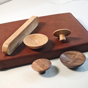 以下が含まれることがあります： さまざまな形状の木製キャビネットノブとプルスのコレクション。ノブにはキノコ型のデザインと丸みを帯びたスタイルがあります。プルは長方形です。木目は各ピースに見え、自然な色合いを示しています。
