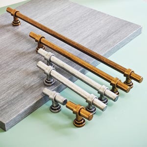 Op de afbeelding: Een assortiment kastgrepen in verschillende maten en afwerkingen. De grepen zijn in brons- en zilverkleuren, met een cilindrisch staafontwerp en een decoratieve basis. De hardware is gerangschikt op een grijze ondergrond.