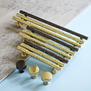 May include: An assortment of cabinet hardware, including gold and black textured bar pulls and matching knobs. The hardware is arranged on a light blue and gray surface, showcasing the different styles and finishes.