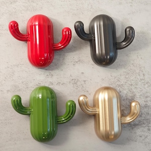 May include: Four cactus-shaped wall hooks in red, dark grey, green, and gold. Each hook has a rounded body with two curved arms extending outwards. The hooks are mounted on a light grey textured wall.
