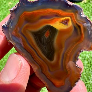 May include: A polished agate stone slice with concentric bands of color. The stone features a dark center that transitions to yellow, orange, and brown hues, with a dark blue edge. The natural stone is held in a person's hand against a green background.