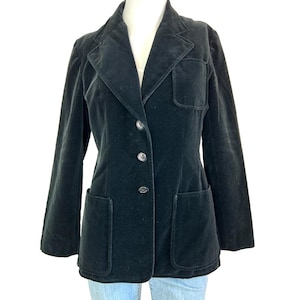 May include: A black velvet blazer featuring a notched lapel, three front buttons, one chest patch pocket, and two lower patch pockets. The jacket has long sleeves and a structured fit, displayed on a mannequin over light blue denim.