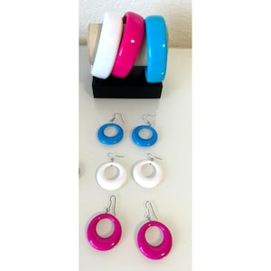 May include: Three thick, smooth bangle bracelets in white, hot pink, and bright blue are displayed on a black stand. Below them are three pairs of matching circular drop earrings in the same vibrant blue, white, and hot pink colors.
