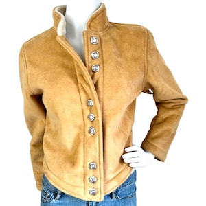May include: A tan suede-textured jacket featuring a high collar lined with white faux shearling. The front closure is secured by a vertical row of eight square, silver-toned metal buttons with decorative detailing. The jacket has long sleeves.