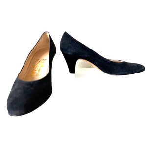 May include: A pair of black suede pumps with a low heel. The shoes have a classic design with a rounded toe and a subtle heel. The interior of one shoe displays the brand name "Ferragamo" in gold lettering.