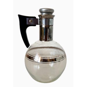 May include: A clear glass pitcher with a round body and a silver metal rim and stopper. The pitcher has a black handle and silver bands around the body. The pitcher is likely for serving beverages.