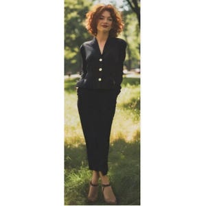 May include: A black skirt suit with a V-neck jacket featuring three gold buttons. The skirt is knee-length. The person is wearing brown sandals. The background is a park with green grass and trees.