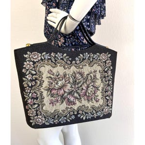 May include: Black tapestry handbag with a floral design in shades of pink, purple, and green. The bag has a rectangular shape with a black border and handles. Gold-toned hardware accents the top.