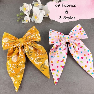 May include: Two fabric hair bows. One bow is mustard yellow with a white floral pattern. The other bow is light pink with a multicolored confetti dot pattern. Text in the image reads 69 Fabrics & 3 Styles. White orchid flowers are visible.
