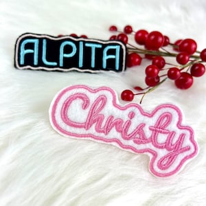 May include: Two embroidered iron-on patches with names. The first patch is black with white outline and says "ALPITA" in blue lettering. The second patch is white with pink outline and says "Christy" in pink lettering.