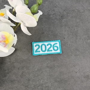 May include: A rectangular patch with the year "2026" embroidered in turquoise thread on a white background. The patch is bordered with a turquoise stitch and is set against a gray background, with white orchid flowers in the upper left corner.