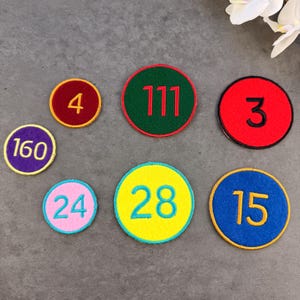 May include: A collection of circular patches with embroidered numbers. The patches feature various color combinations, including red, green, yellow, blue, and purple, with numbers like 4, 3, 15, 24, 28, 111, and 160. Each patch has a contrasting border.
