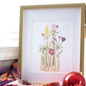 May include: A framed print of a floral arrangement in a gold frame. The flowers are in various shades of pink, purple, and yellow. The flowers are arranged in a vase made of clear glass tubes.