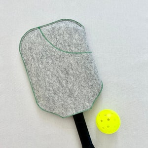 May include: A gray felt pickleball paddle with a green stitched design and a black handle. A yellow pickleball is next to the paddle.