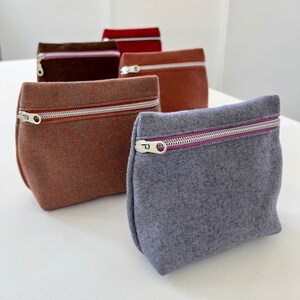 May include: Five felt zipper pouches in different colors, including brown, orange, gray, and red. The pouches are lined up in a row on a white surface.